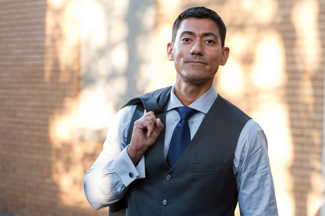 Rocky Vega in a professional portrait, gray vest and tie, in front of a brick wall.